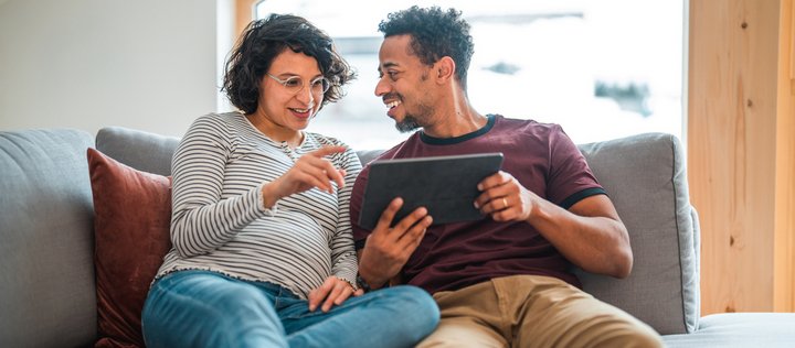 Schwangere und Mann lächelnd auf Sofa im Gespräch mit Laptop