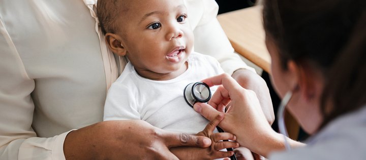 Ein Baby sitzt auf dem Schoß der Mutter. Eine Kinderärztin untersucht das Baby mit einem Stethoskop.