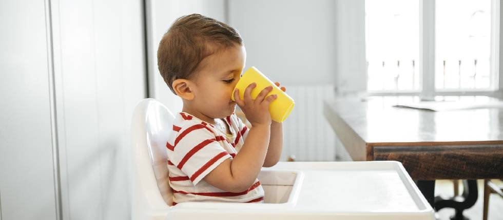 Kleiner Junge sitzt in Hochstuhl und trinkt Wasser aus einem gelben Becher.
