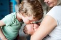Blonder Junge küsst sein Geschwisterkind im Säuglingsalter auf die Stirn. Die Mutter hält das Baby im Arm. 