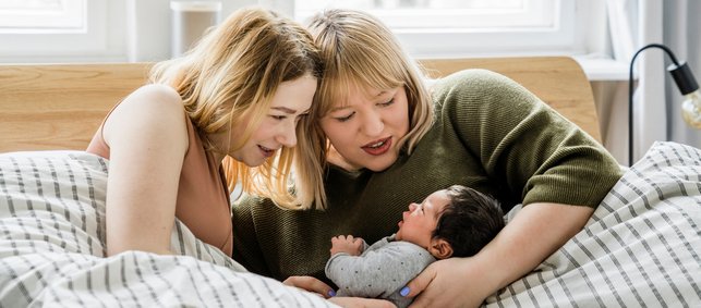 Zwei Frauen liegen mit einem kleinen Baby im Bett und schauen es verbunden an