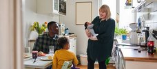 In einer Küche sitzen ein Mann und ein Kind am Tisch, eine Frau mit Baby im Arm steht ihnen zugewandt daneben.