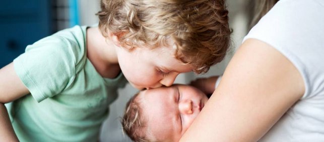 Blonder Junge küsst sein Geschwisterkind im Säuglingsalter auf die Stirn. Die Mutter hält das Baby im Arm. 