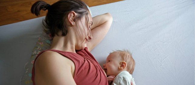 Frau und Baby liegen seitlich einander zugewandt auf einer Matraze. Die Frau stillt das Baby.